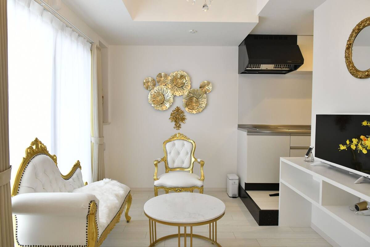 A luxurious room with a white sofa, a gold-trimmed armchair, a round gold table, and a TV on a white stand.