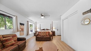 Une pièce spacieuse avec un grand lit, un canapé et une table ronde. On y trouve un ventilateur de plafond, une fenêtre avec des rideaux et une porte dotée d’un miroir.
