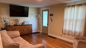 A living room with a wooden floor, beige sofas, a TV mounted on the wall, a wooden cabinet, and a window with curtains. - The Family Escape Ranch (Erie)