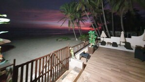 Eine Holzterrasse mit Blick auf einen Strand, Palmen und einen Sonnenuntergang.