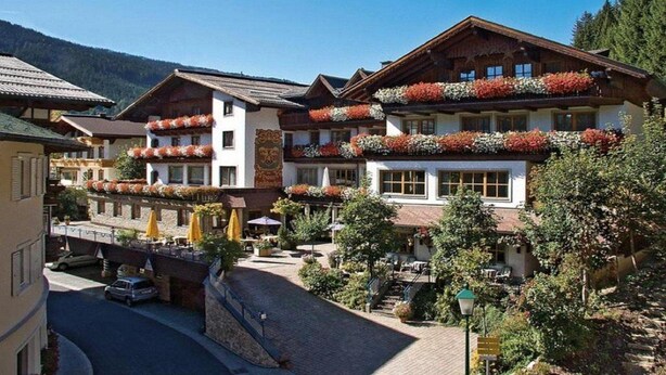 Un hotel alpino tradicional con múltiples balcones decorados con flores, un patio empedrado y un paisaje montañoso de fondo.