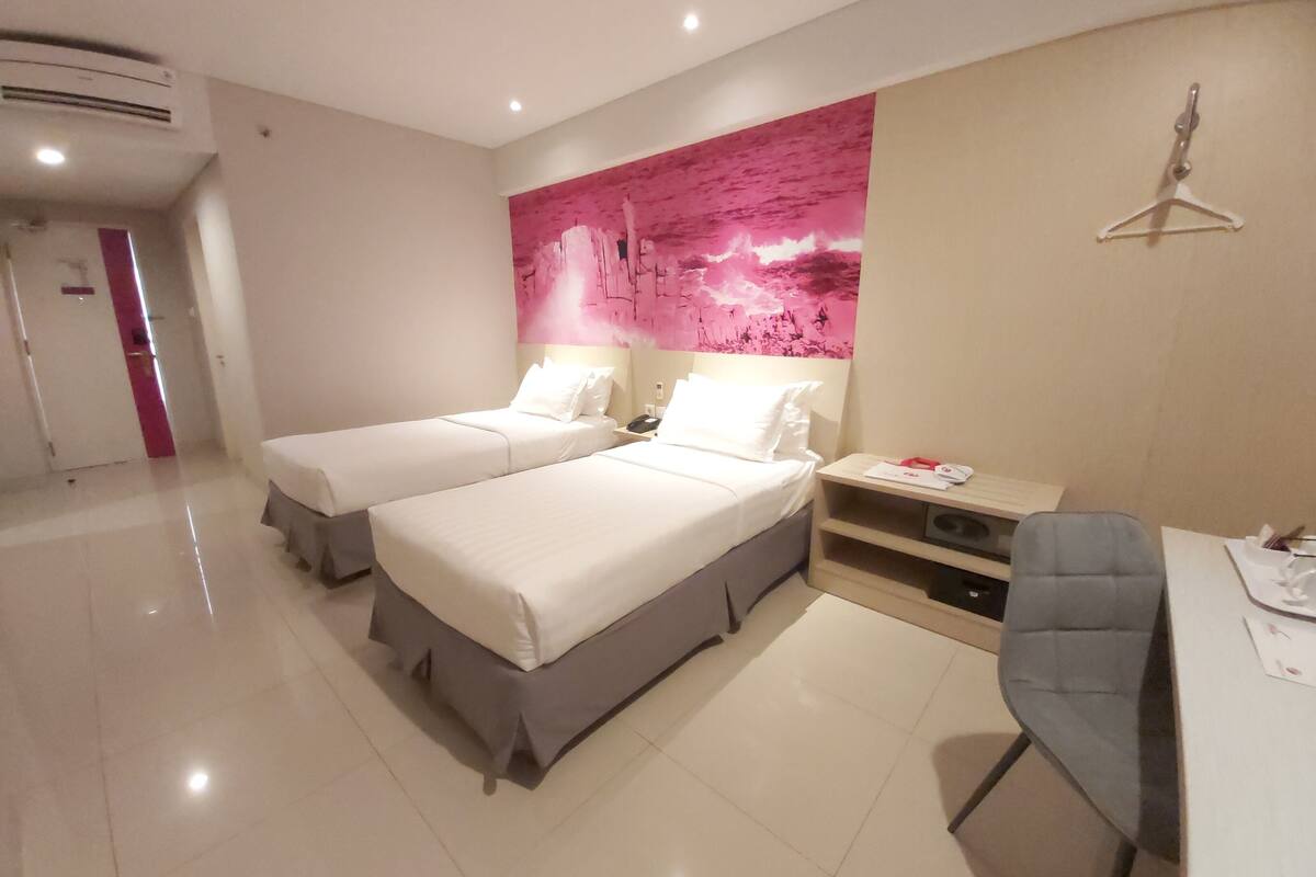 A hotel room with two beds, a desk with a chair, a large pink abstract painting on the wall, and a hanger on the wall.