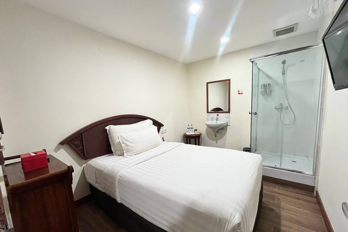 A hotel room with a bed, a bedside table, a small table with toiletries, a sink, and a shower.