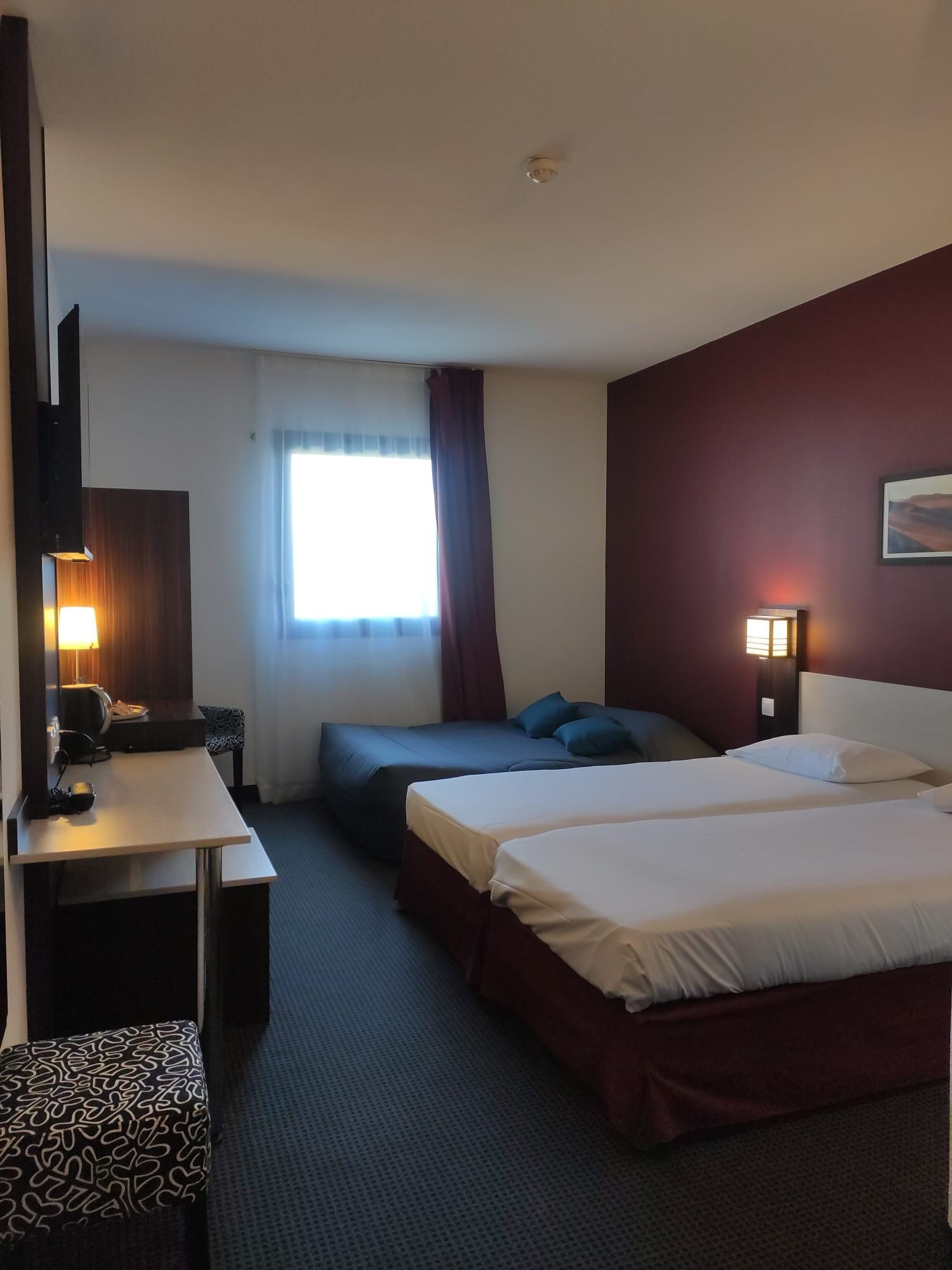 A hotel room with a bed, a desk, a chair, a TV, and a window with curtains.
