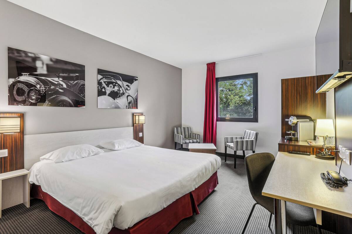 Hotelkamer met een groot bed, een bureau met telefoon, twee stoelen, een raam met rode gordijnen en twee wandlampen.