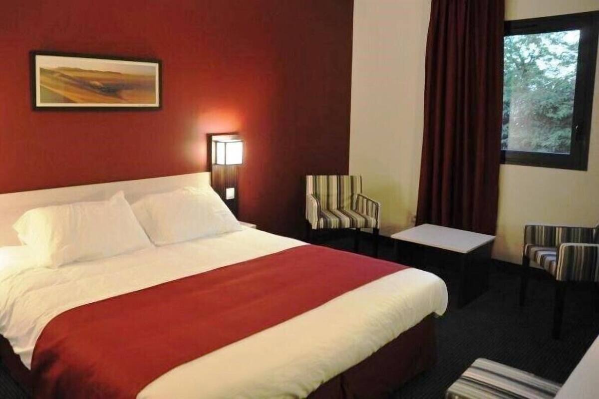 Een hotelkamer met een groot bed, twee nachtkastjes, een stoel, een klein tafeltje en een schilderij aan de muur.