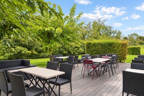 Une terrasse extérieure aménagée avec des meubles et des tables en osier, entourée de verdure et d’arbres.
