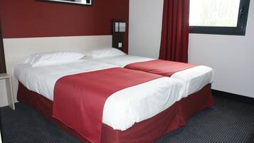 A double bed with white and red bedding, a small bedside table, a nightstand, a red curtain, and a window with a view of trees.