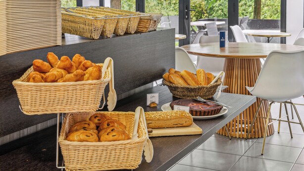 Un buffet de petit-déjeuner avec des paniers en osier remplis de viennoiseries et de pain, une table en bois et des chaises blanches.