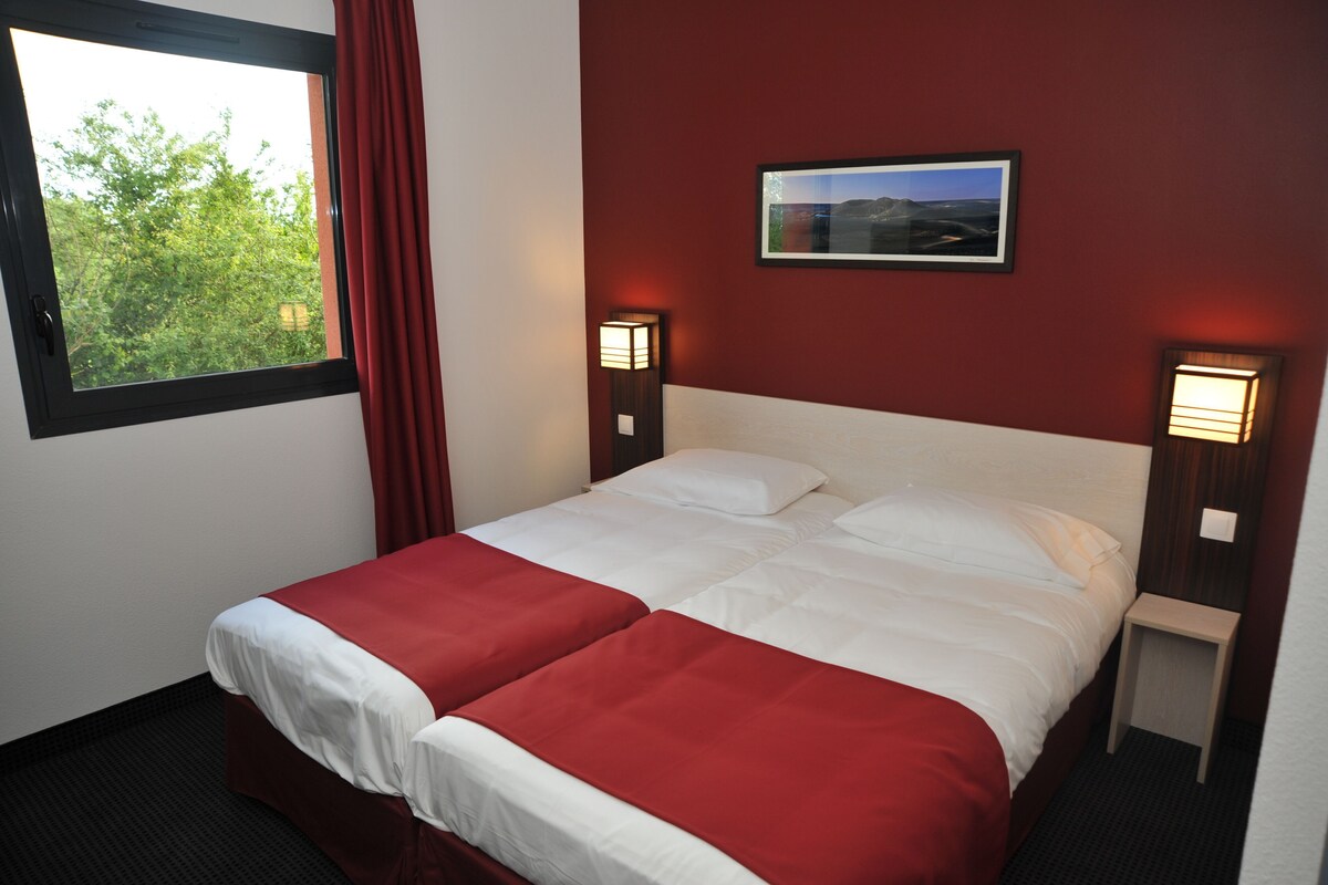 A double bed with white and red bedding, two bedside lamps, a framed picture on the wall, and a window with a view of trees.