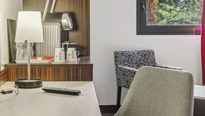 Une chambre d’hôtel équipée d’un bureau, d’une lampe, d’une chaise, d’un radiateur et d’une fenêtre donnant sur des arbres.