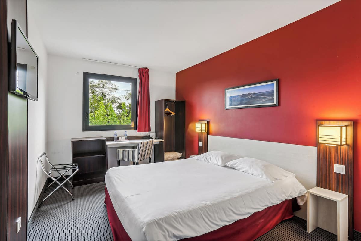 Hotelkamer met een bed, bureau, stoel, televisie en raam met uitzicht op groen.