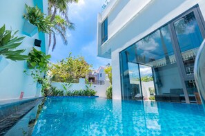 A modern pool area with a clear blue pool, surrounded by lush greenery and a white building with large windows.