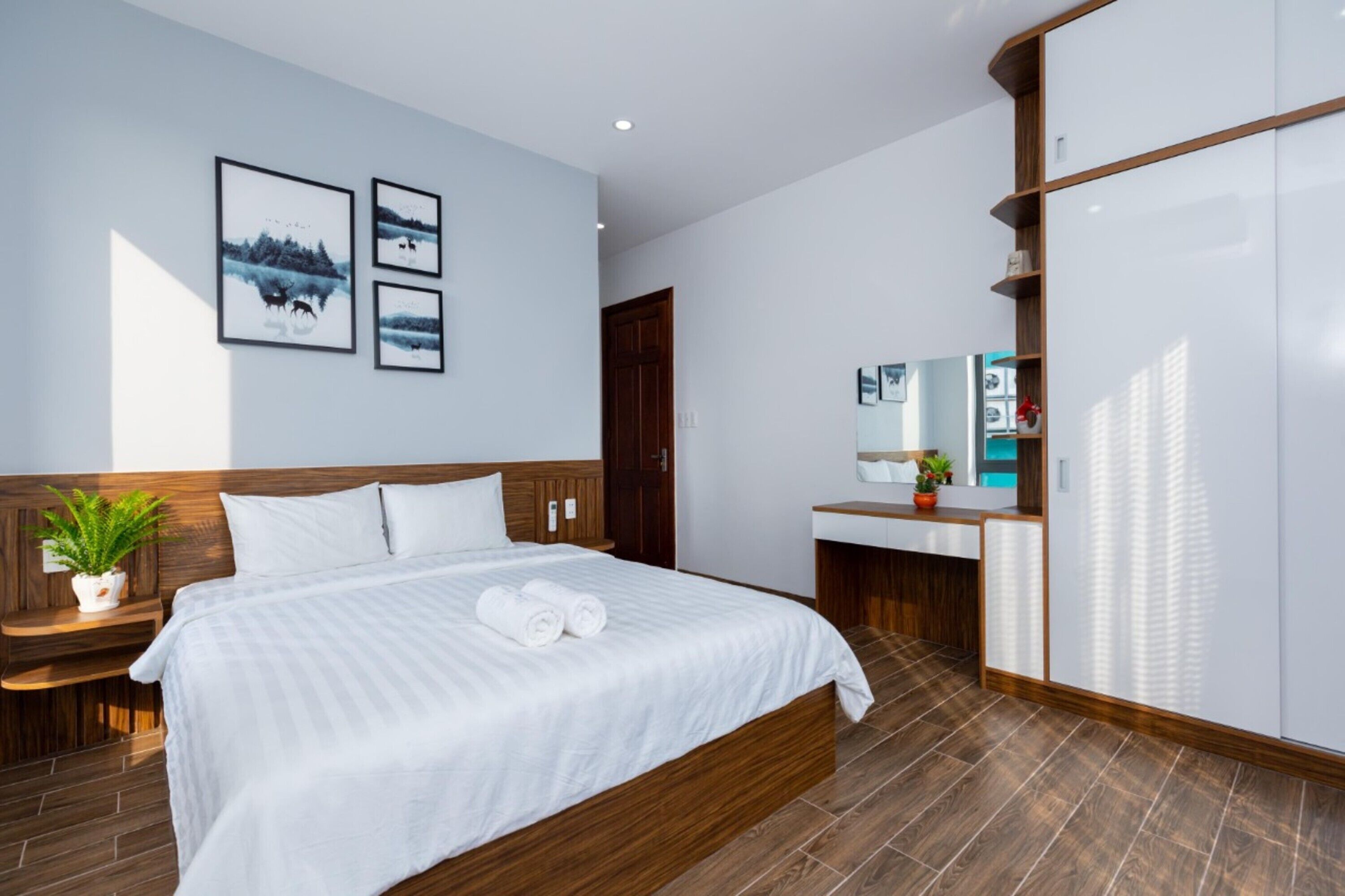 A modern hotel room with a large bed, wooden headboard, a bedside table with a plant, a built-in wardrobe, and a bathroom with a mirror and sink.