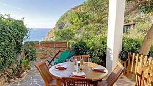 Mesa de madera lista para comer con vista al mar, rodeada de follaje y un muro de piedra.