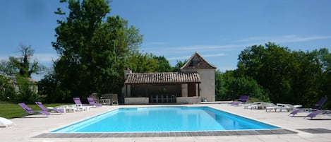 A rectangular swimming pool with a tiled deck, surrounded by lounge chairs and a small building with a tiled roof.