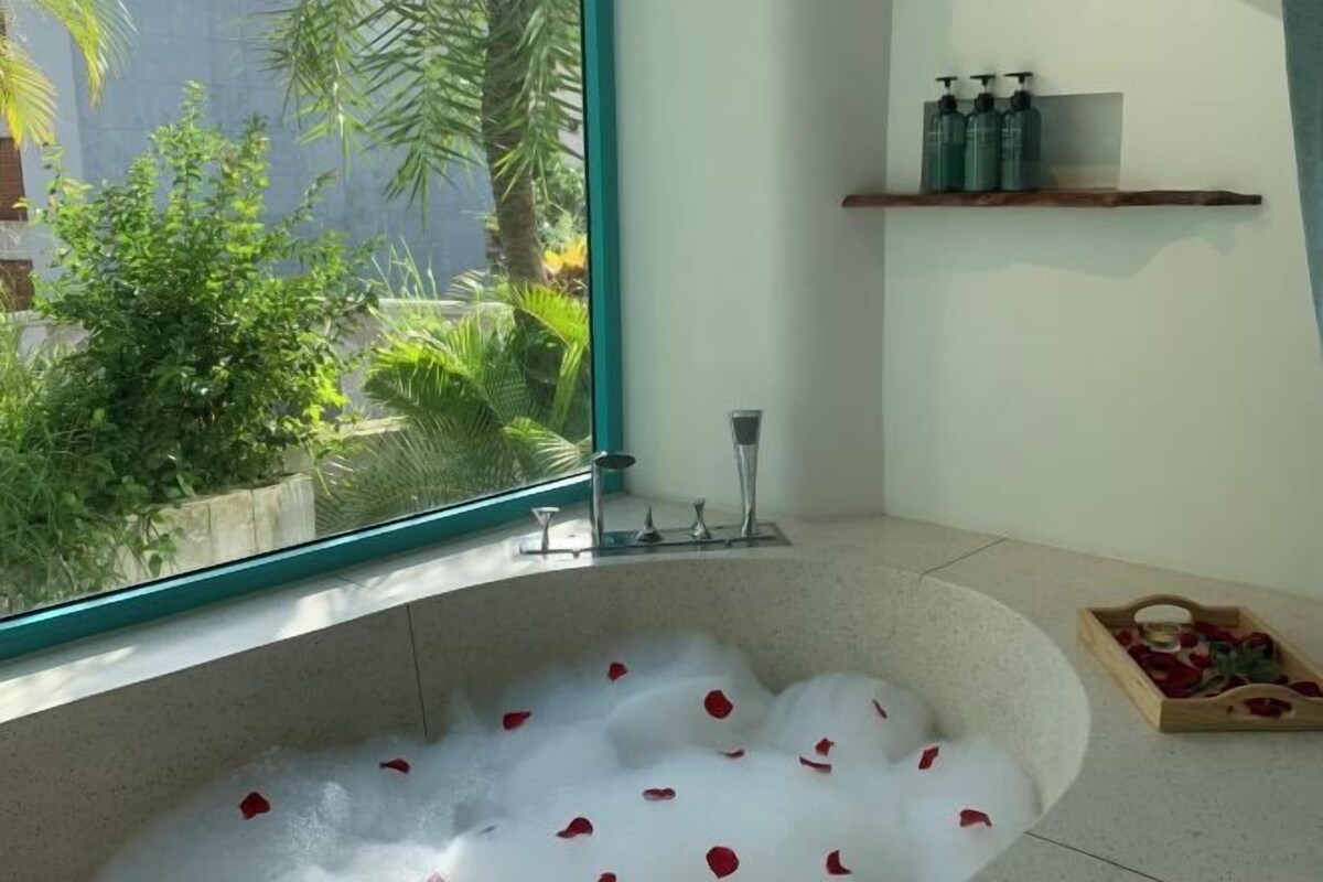 A bathtub with rose petals, a window with a view of greenery, and a shelf with toiletries.