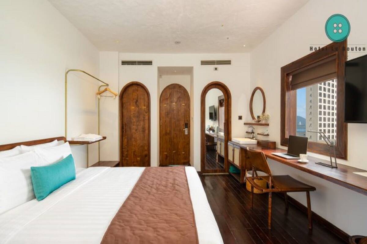 Une chambre d’hôtel avec un lit, un bureau et une chaise, un miroir et une télévision. Il y a trois portes en bois et une fenêtre donnant sur un paysage agréable.