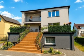 A two-story residential building with a staircase leading to a balcony, a small garden area, and a visible door.