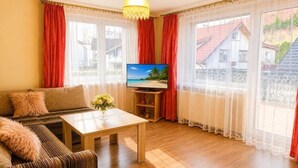 A furnished room with a sofa, a coffee table, a TV, and large windows with curtains. - Holiday Home near Baltic Sea & Sandy Beaches (Miedzyzdroje)