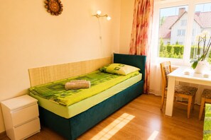 Un lit simple avec une tête de lit verte et une couverture assortie, une table de chevet, une petite commode blanche, une lampe fixée au mur, une fenêtre avec des rideaux et une vue sur un bâtiment à l’extérieur.