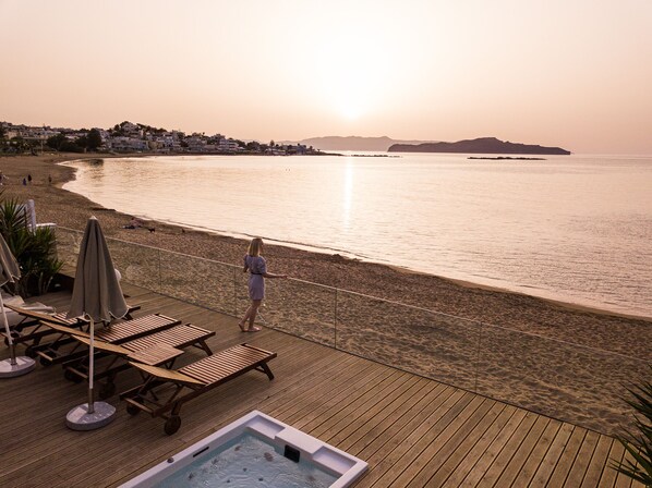 Une terrasse en bois avec des chaises longues et un jacuzzi, donnant sur une plage où l’on peut admirer le coucher du soleil.