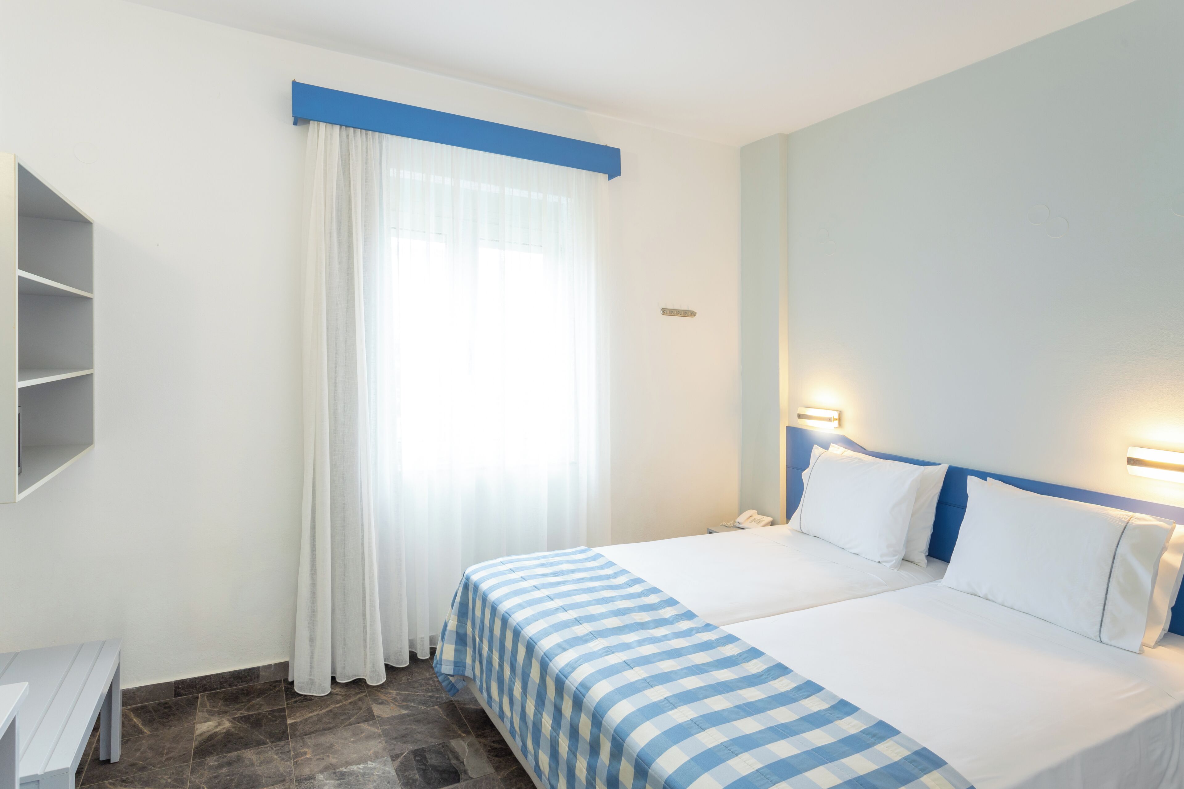 A hotel room with a bed, a bedside table, a nightstand, a window with curtains, and a wall-mounted light.