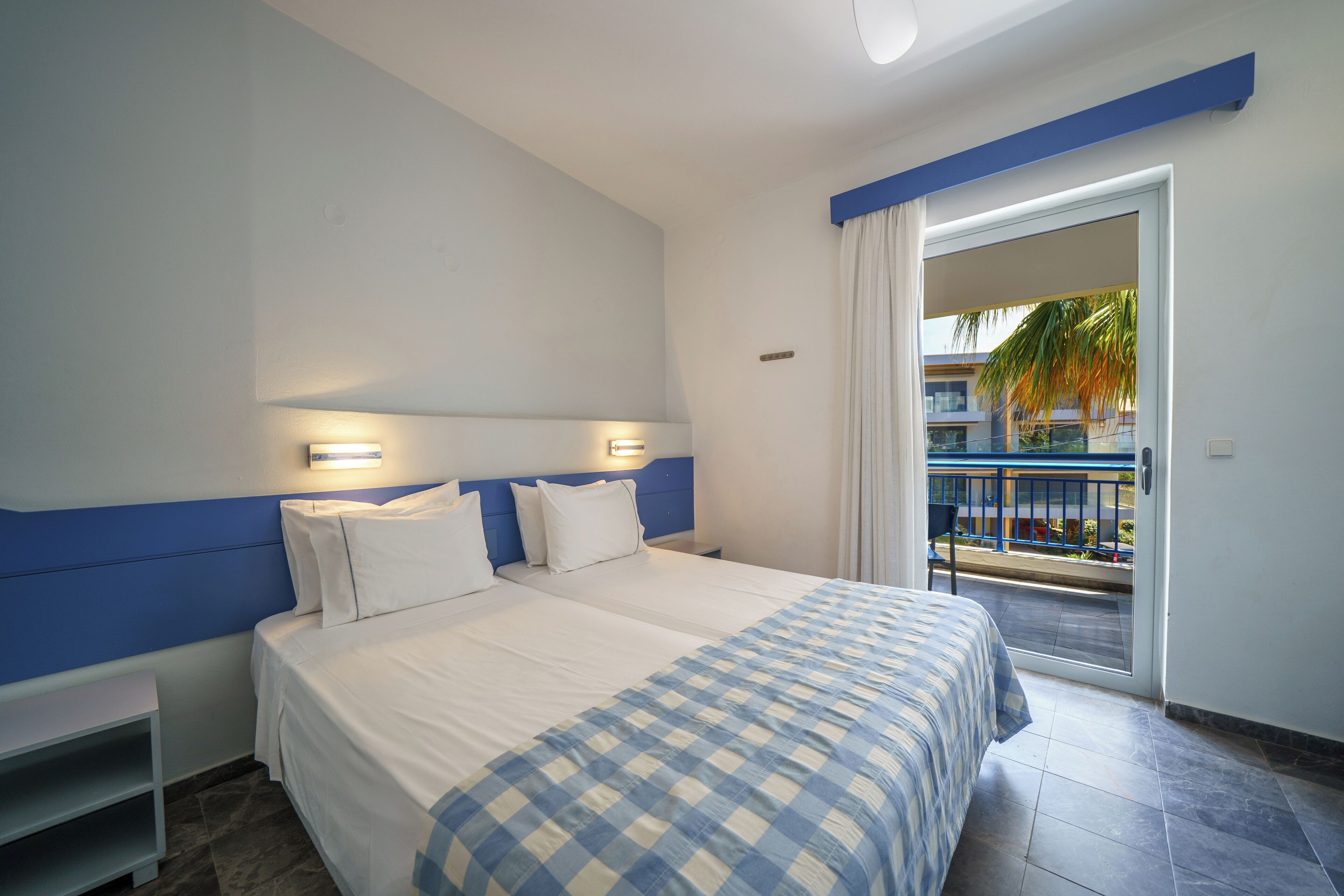 A hotel room with a bed, nightstands, a window with curtains, and a view of a balcony with a palm tree.