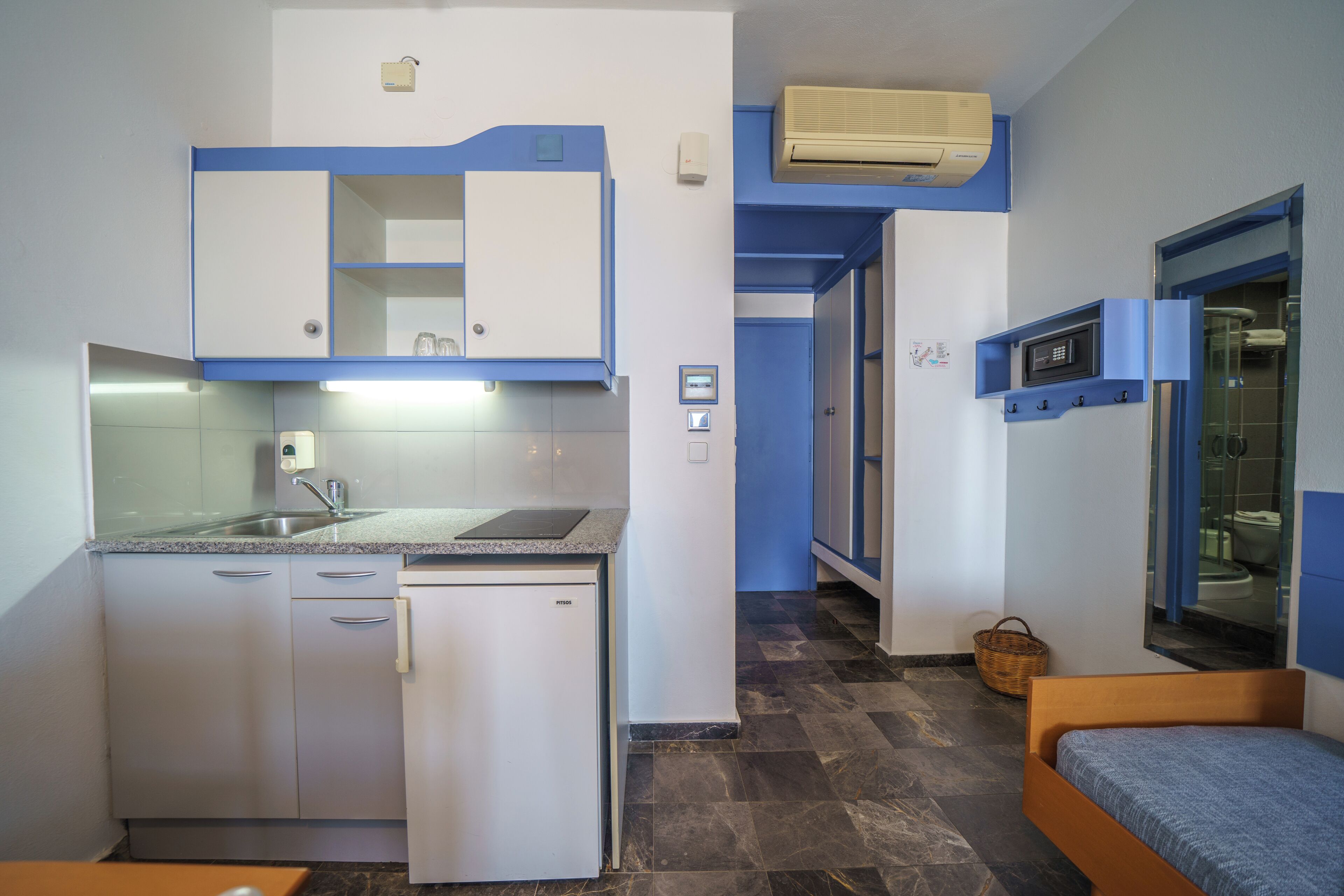 A compact kitchen with a refrigerator, sink, and microwave. There is a small bed with a blue mattress and a wooden headboard.