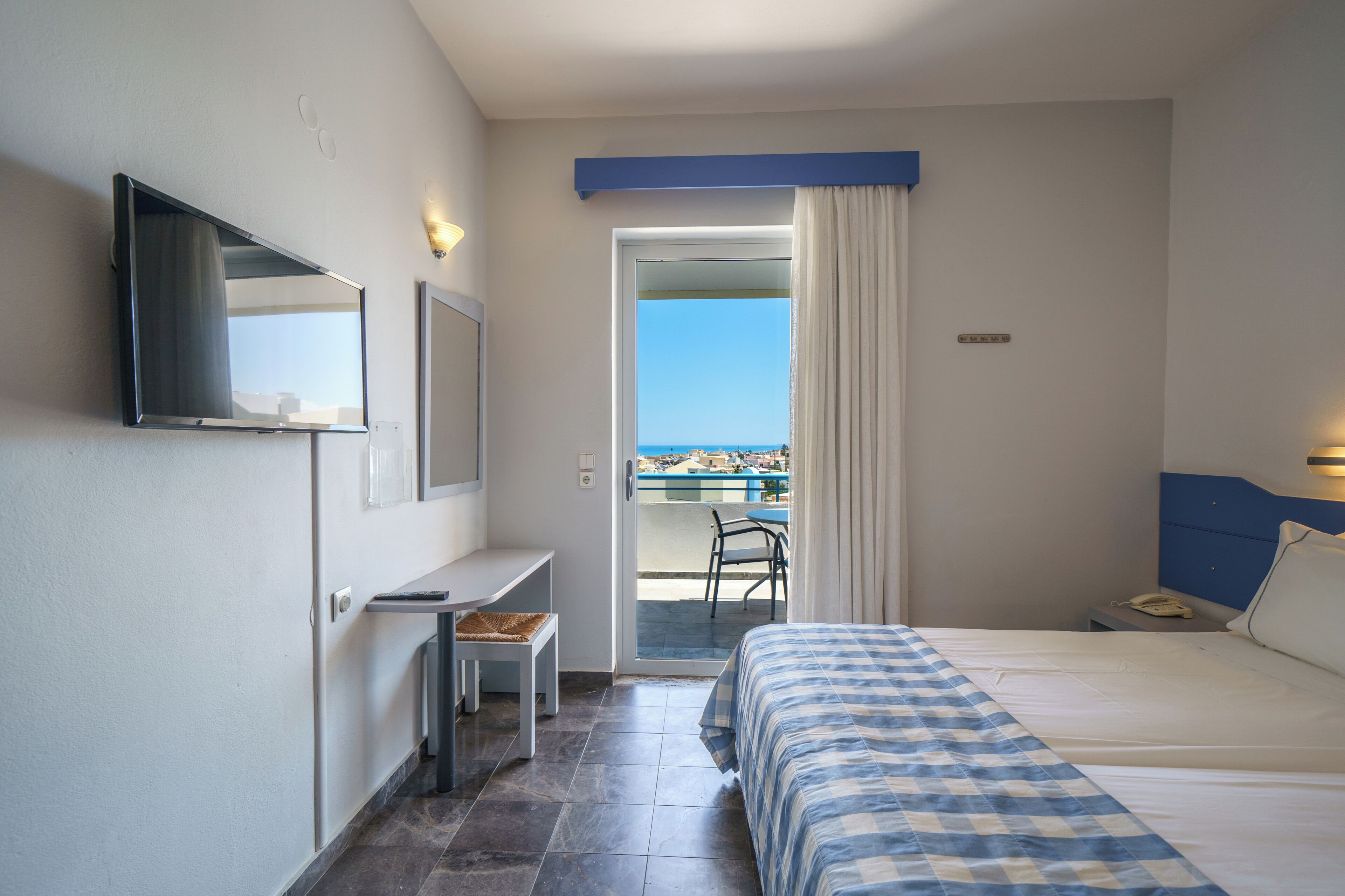 A hotel room with a bed, a desk, a chair, a TV, and a view of the sea.