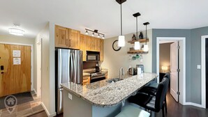 Une cuisine moderne dotée de plans de travail en granit, d’appareils électroménagers en acier inoxydable, d’armoires en bois et de luminaires suspendus.