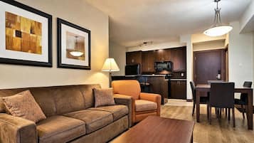 A living room with a brown leather sofa, a wooden coffee table, and a dining area with a dark wooden table and chairs. There is a kitchen with dark wooden cabinets and a microwave.