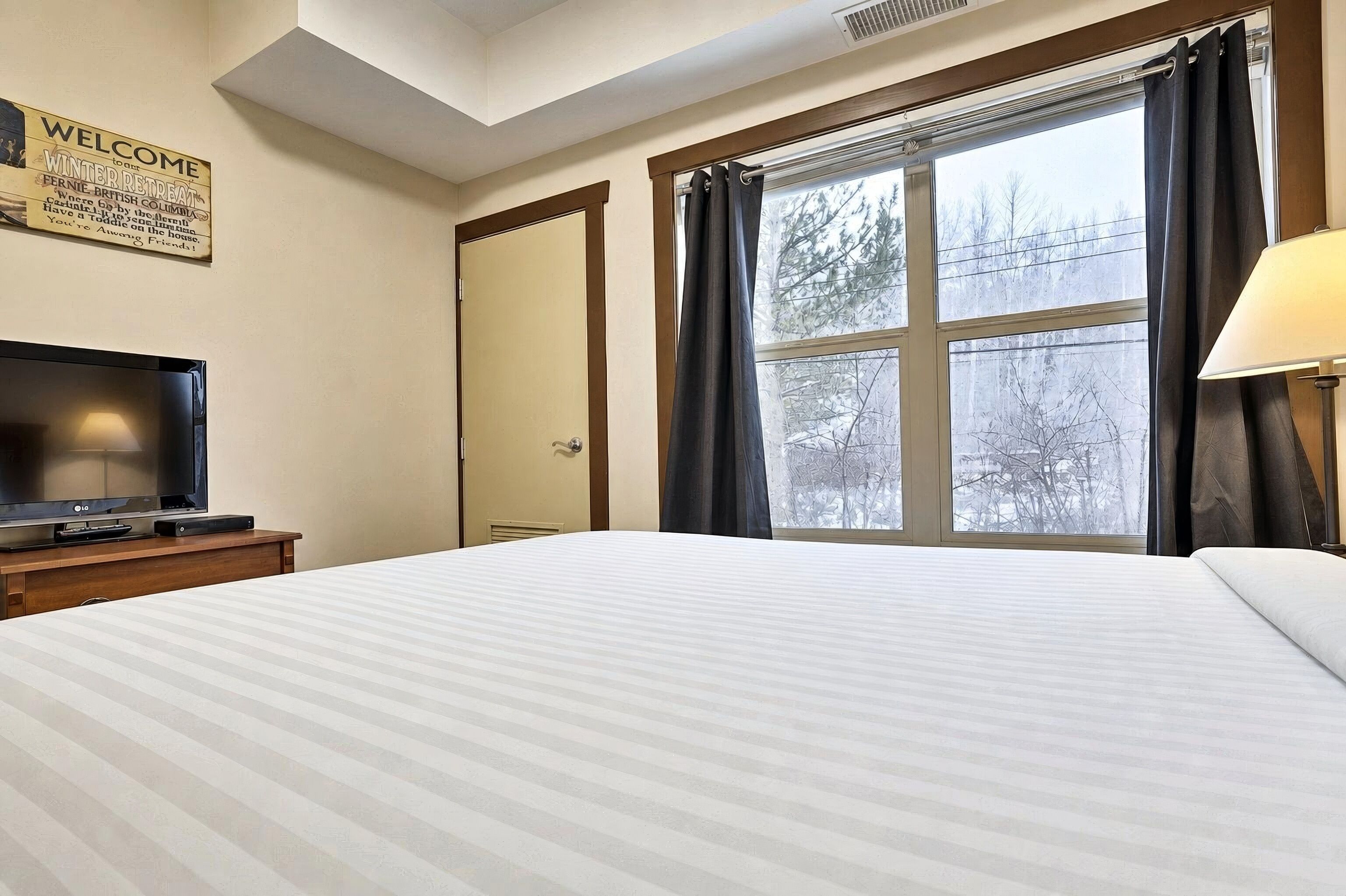 Quarto de hotel com uma cama, uma televisão em um suporte, uma janela com cortinas e um letreiro de boas-vindas na parede.