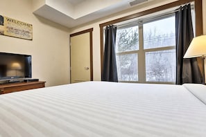 A hotel room with a bed, a television on a stand, a window with curtains, and a welcome sign on the wall. - 1 BDRM / King / AC / Hot Tub & Pool (Fernie)