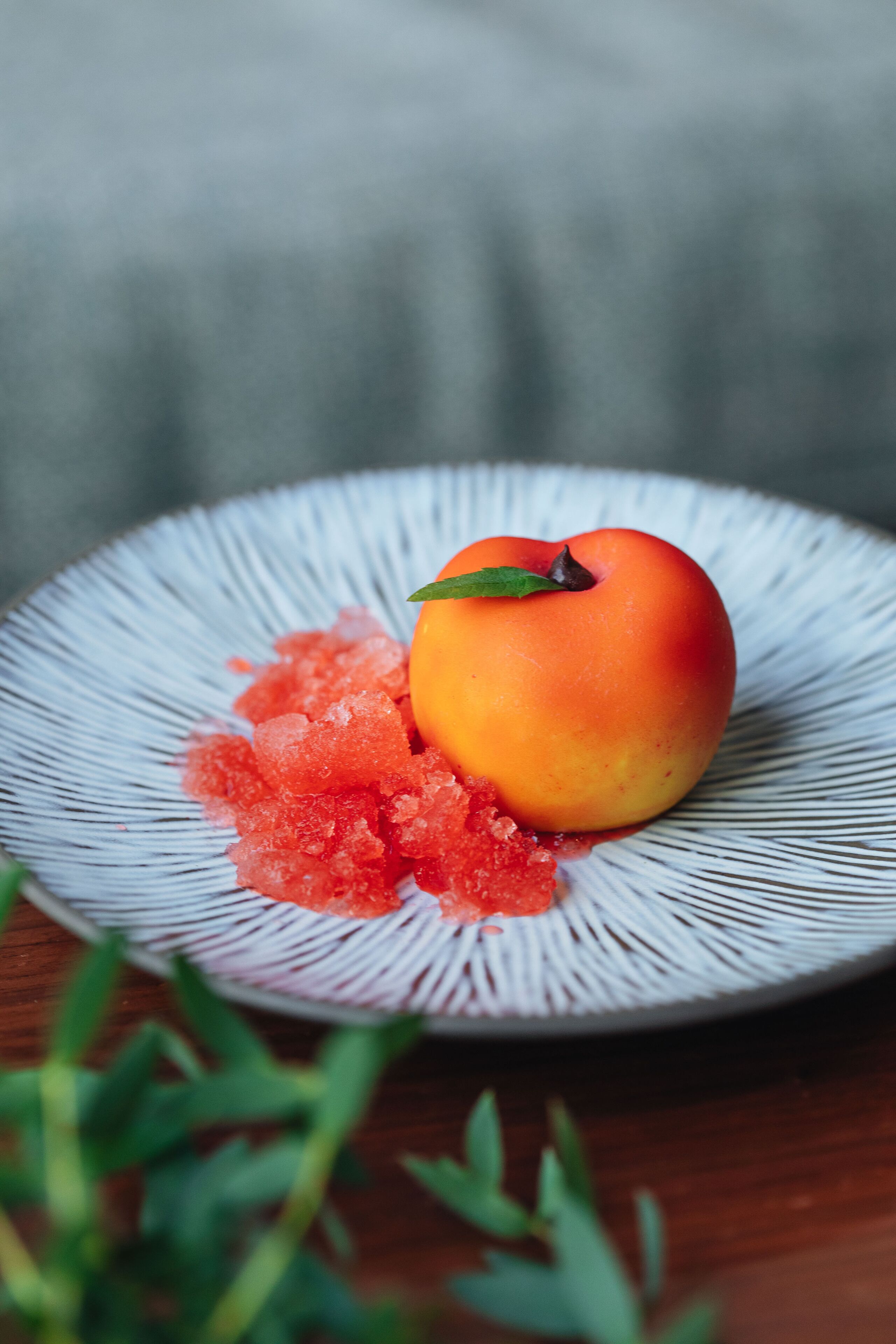 A single peach on a plate with a textured white rim, accompanied by a small pile of red granules, possibly sugar, on a wooden surface.