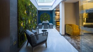 Et moderne interiør med et glasloft, en grøn væg med planter, et siddeområde med sofa og stole, samt en gul lænestol.