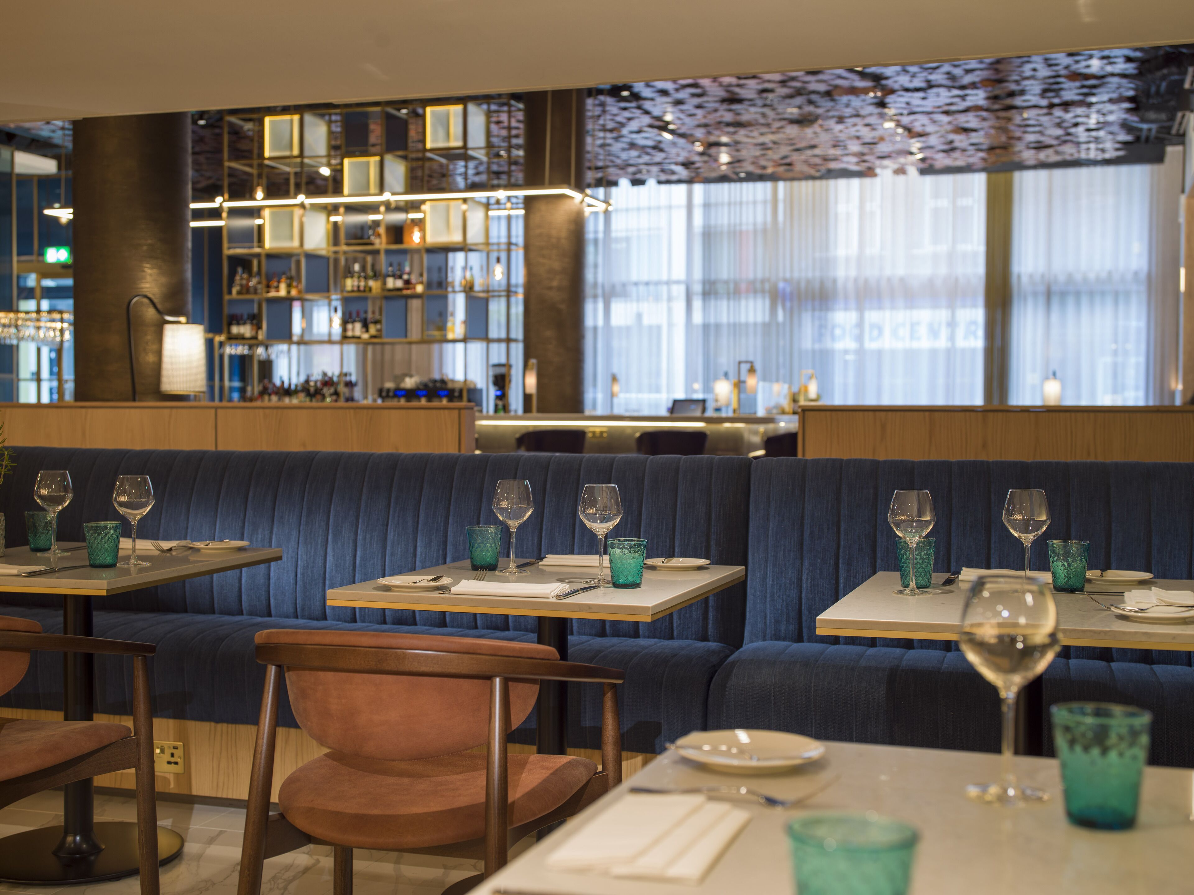 A restaurant with booths and tables, some with blue cushions, and a bar area in the background.