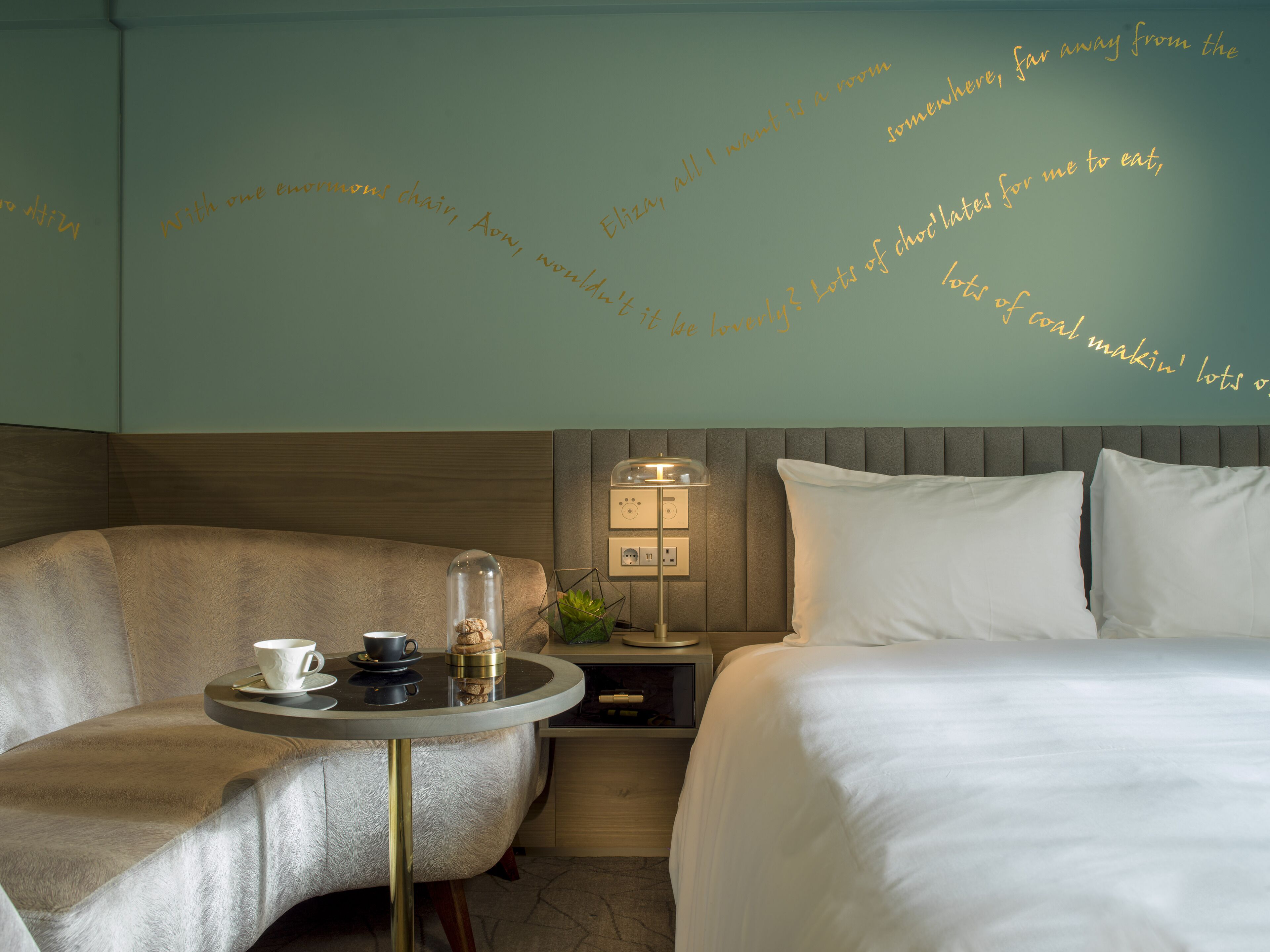 A hotel room with a bed, a chair, a table with a cup and saucer, and a lamp.