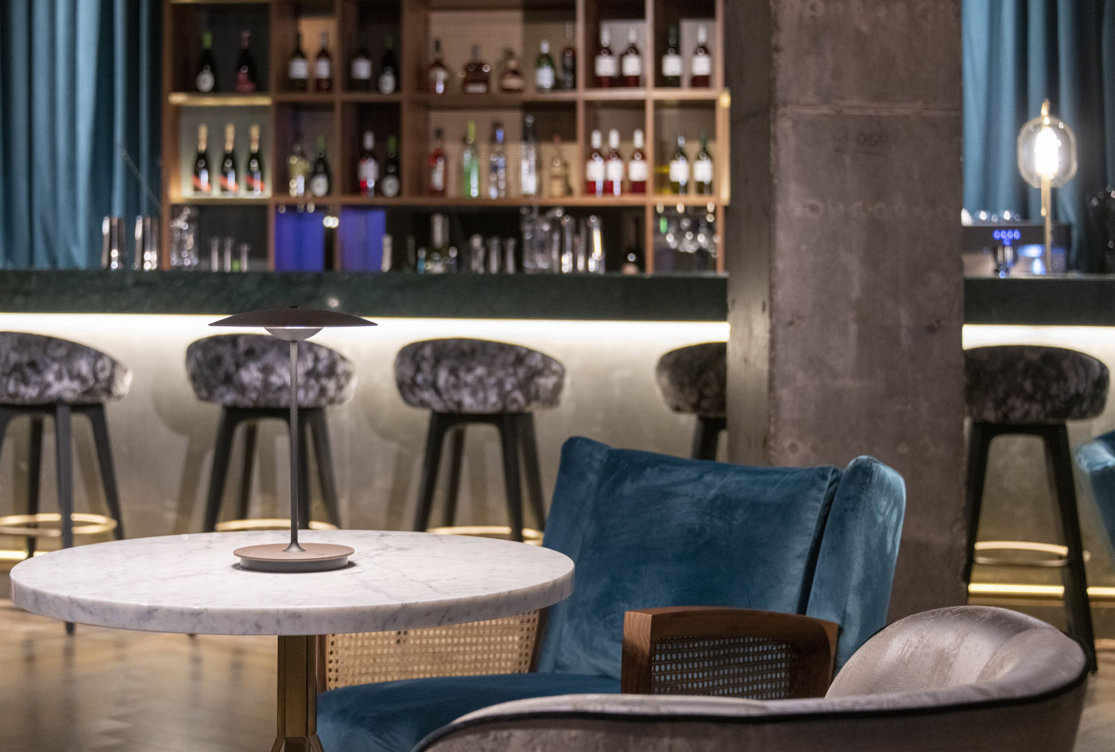 A bar area with a marble table, blue upholstered chairs, and a variety of bottles on shelves.