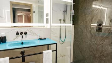 Casa de banho moderna com cabine de chuveiro em vidro, parede revestida a mármore e lavatório azul com bancada de vidro.