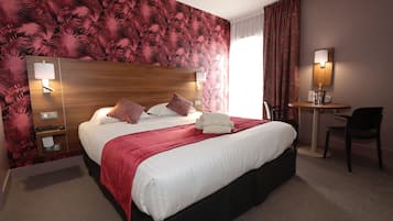 A hotel room with a large bed, a desk, a chair, and a wall-mounted lamp.