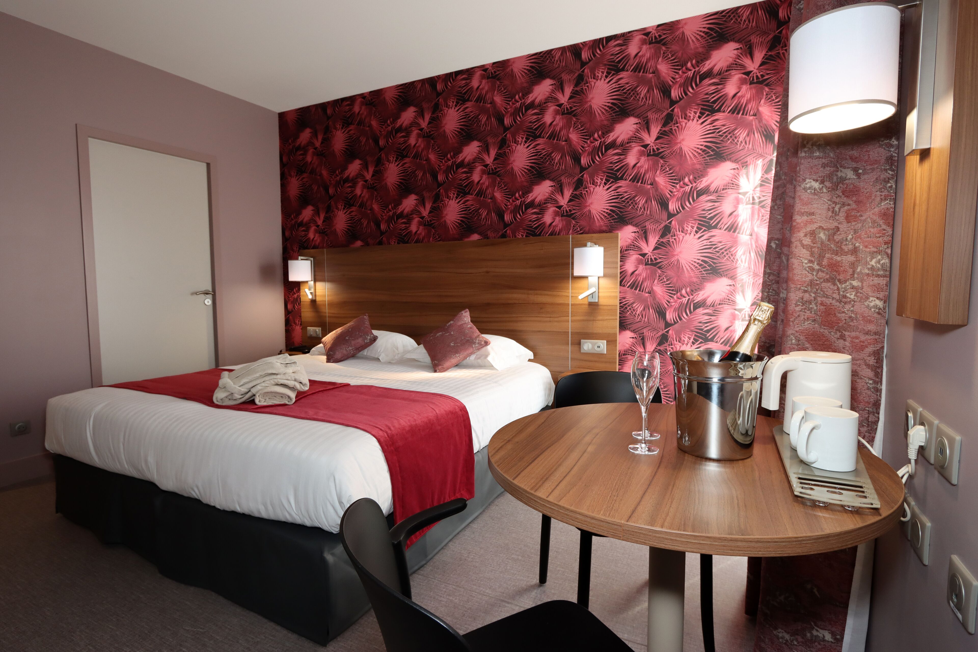 Une chambre d’hôtel avec un lit, une table en bois sur laquelle se trouvent une carafe et des tasses, et un mur décoré d’un motif rouge et noir.