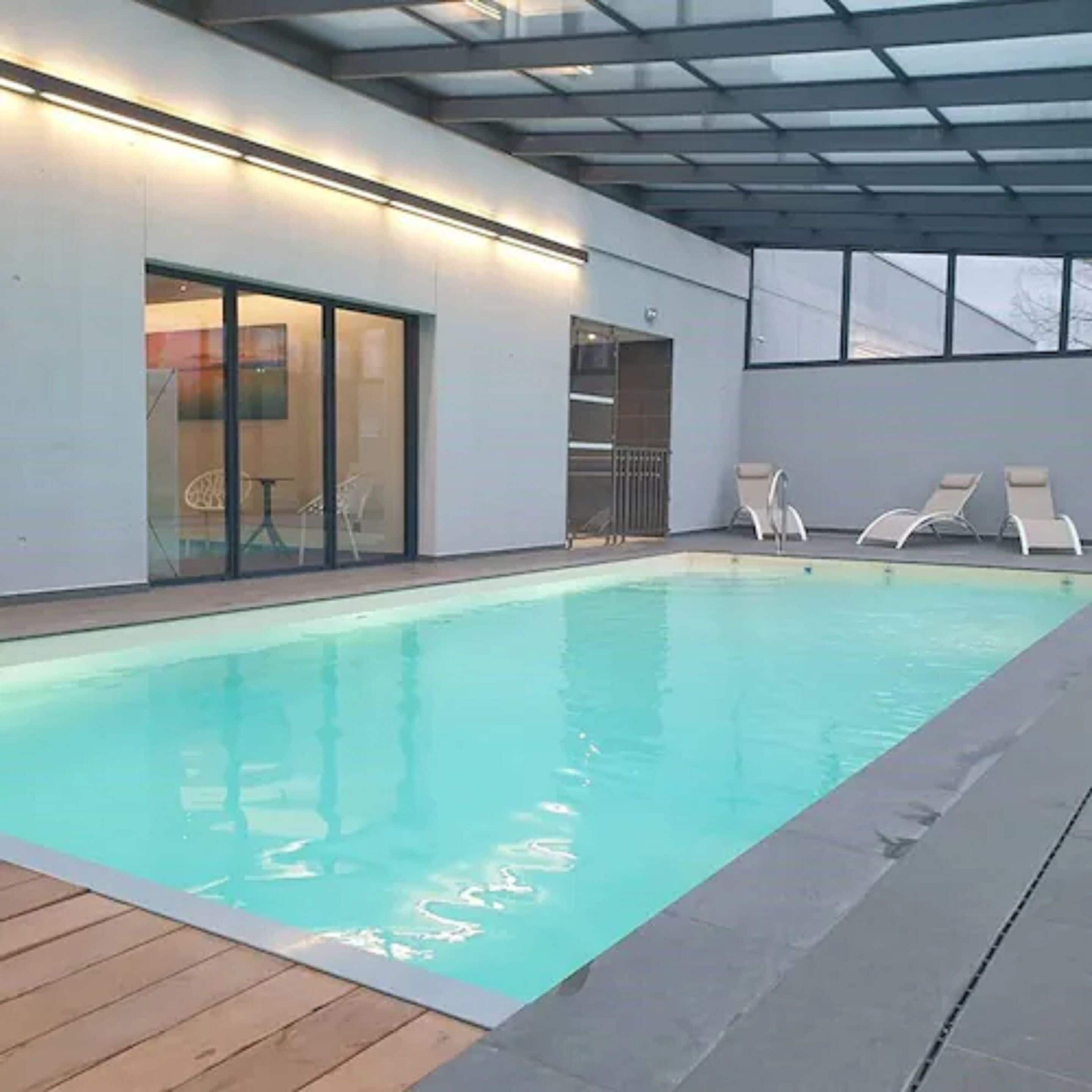 Una piscina moderna al chiuso con acqua cristallina, circondata da un pontile in legno e poltrone da relax bianche.