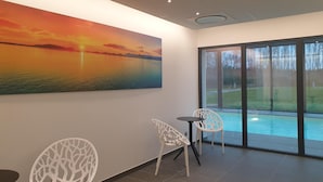 Une pièce moderne avec un grand tableau représentant un coucher de soleil sur l’eau, deux chaises blanches, une petite table et une vue sur une piscine, visible à travers de grandes portes vitrées.