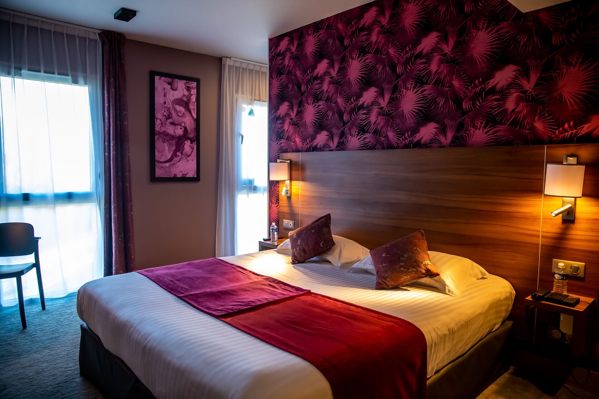 A hotel room with a large bed, two bedside lamps, a nightstand, a chair, and a wall-mounted artwork.