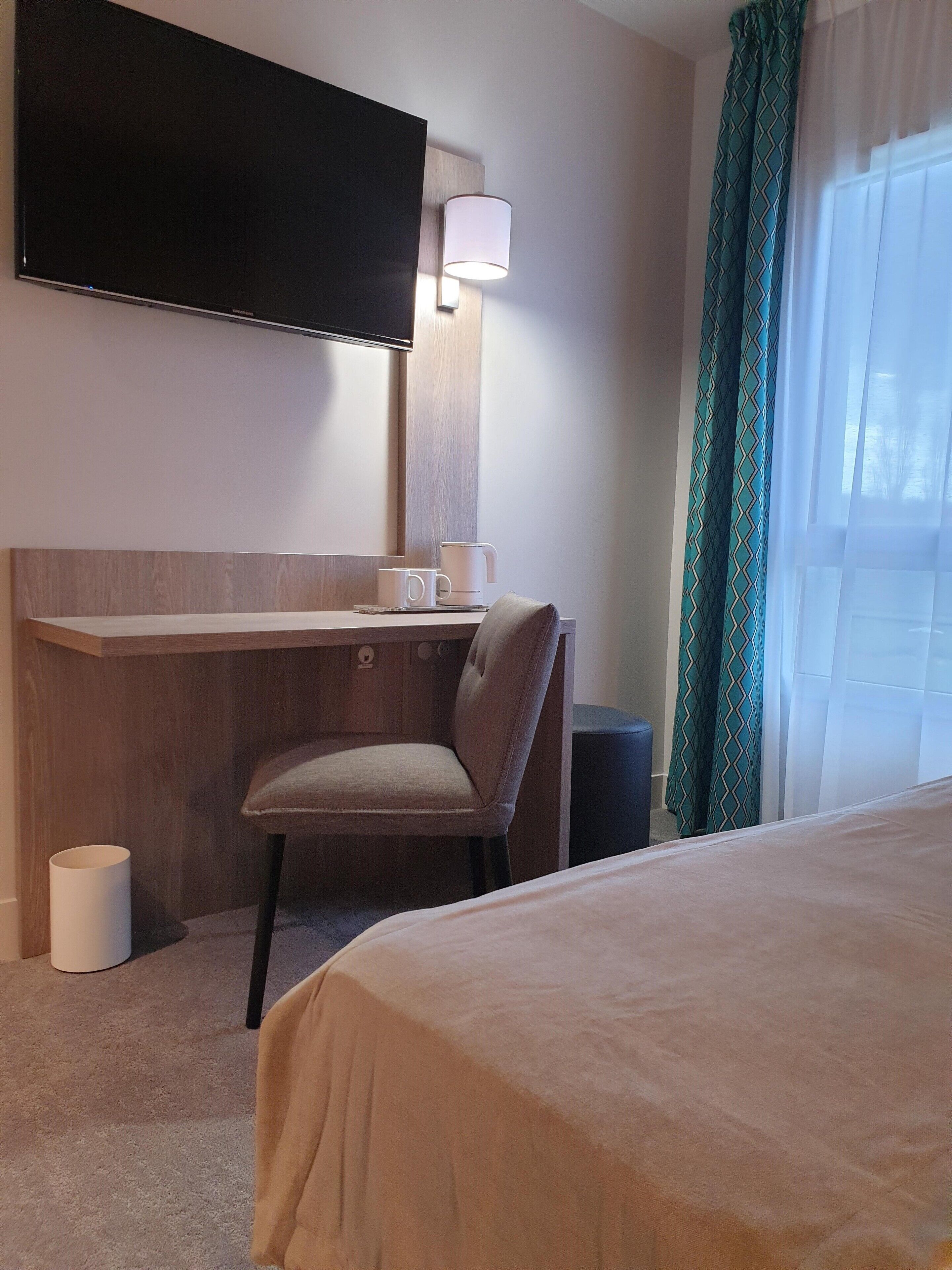 Quarto de hotel com cama, escrivaninha com cadeira, televisão na parede e janela com cortinas.