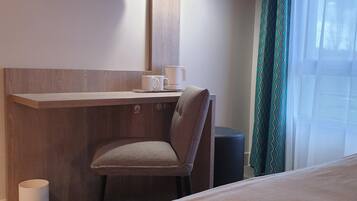 Quarto de hotel com cama, escrivaninha com cadeira, televisão na parede e janela com cortinas.
