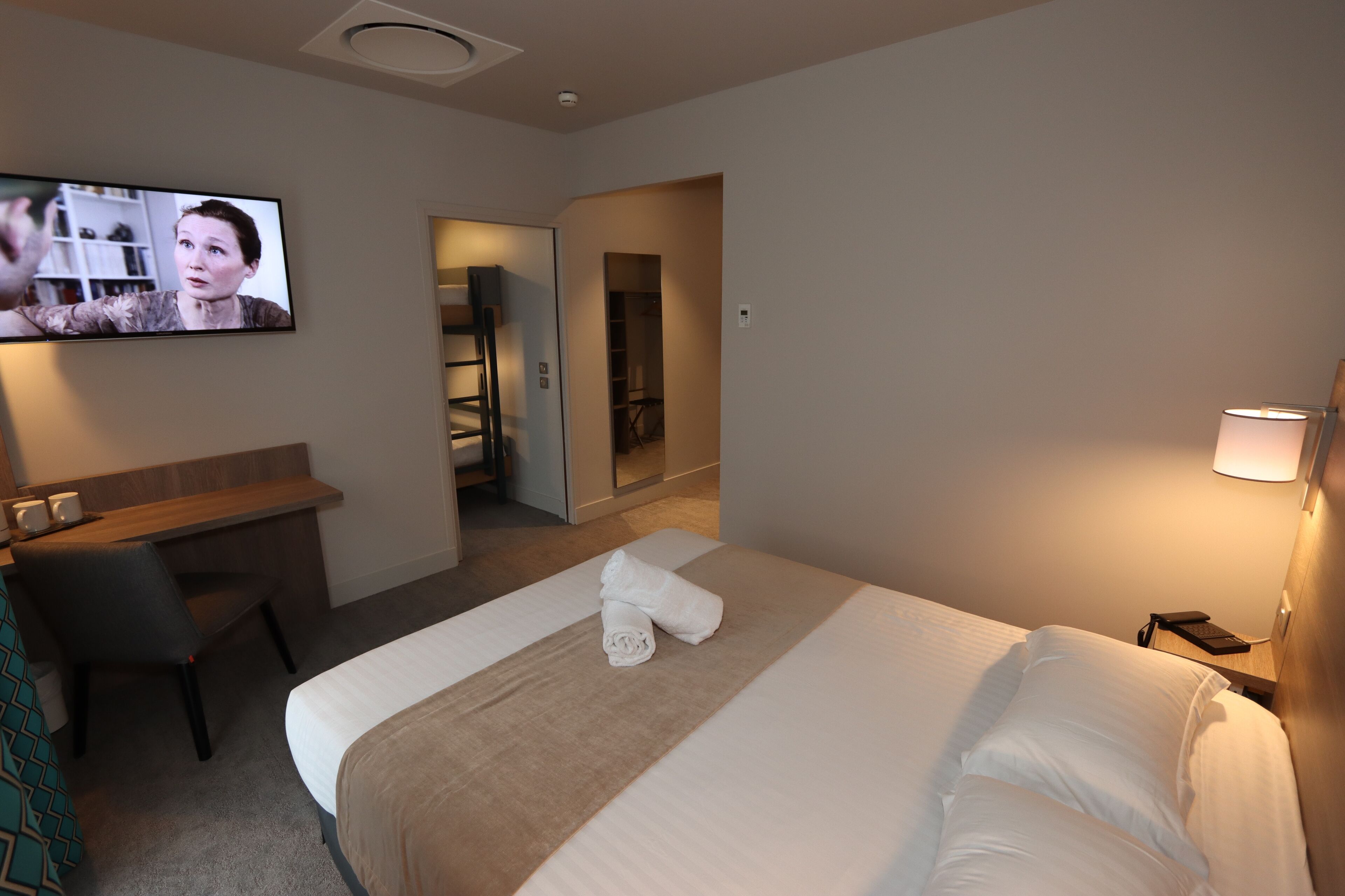 A hotel room with a bed, a desk, a chair, a TV, and a lamp.