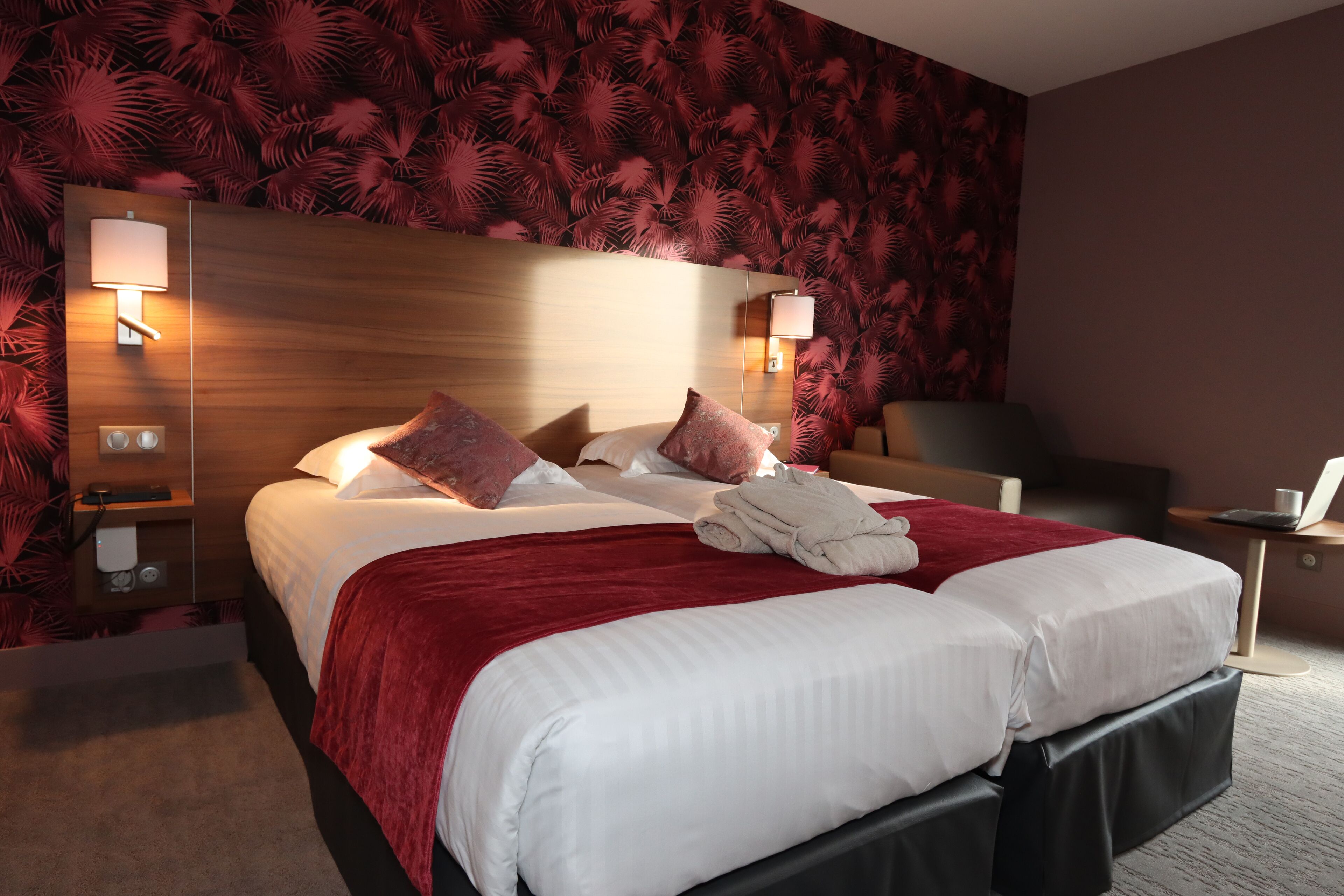 A hotel room with a large bed, two bedside lamps, a red throw blanket, and a neatly folded towel on the bed.