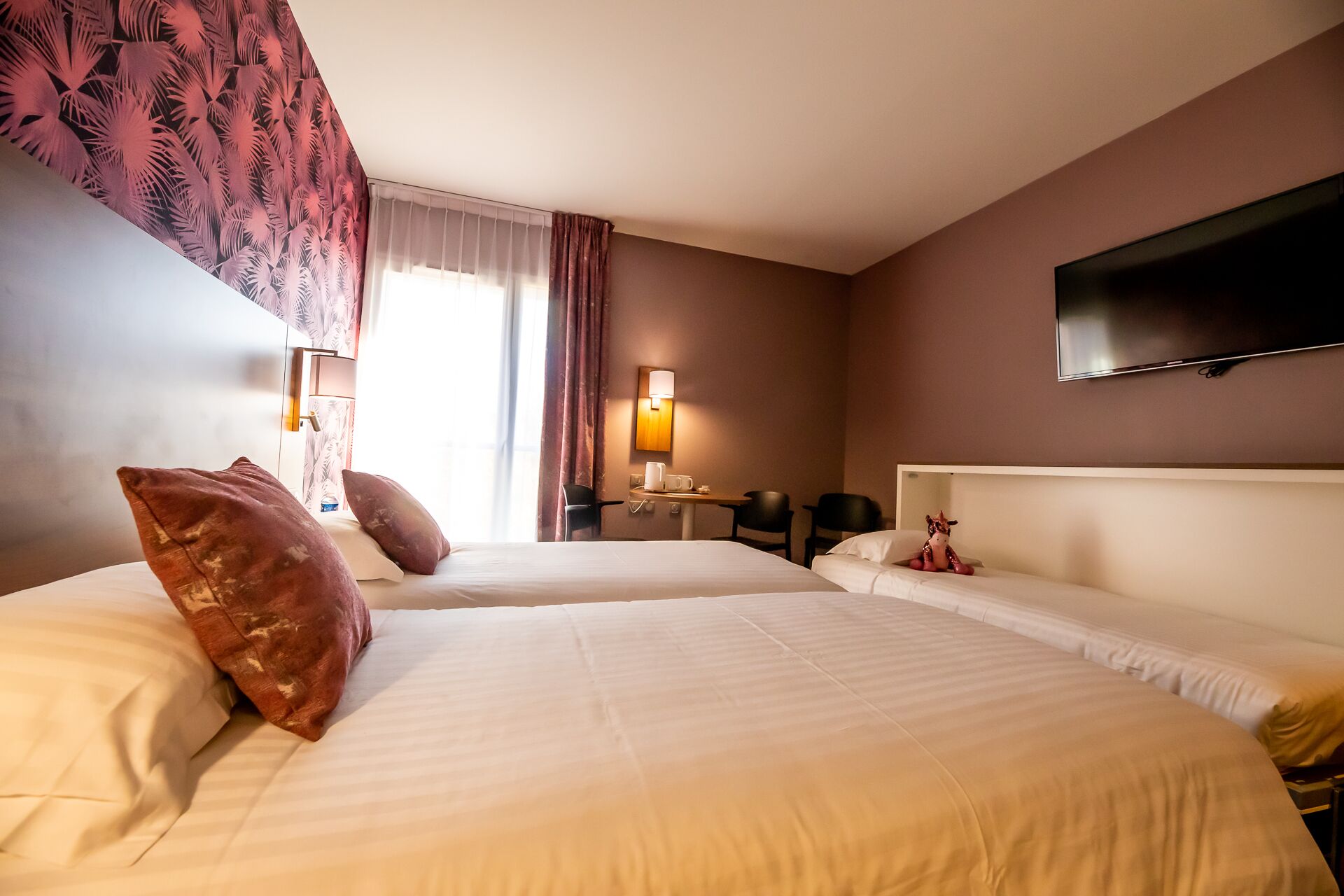 A hotel room with two beds, a TV mounted on the wall, a desk with a chair, and a window with curtains.